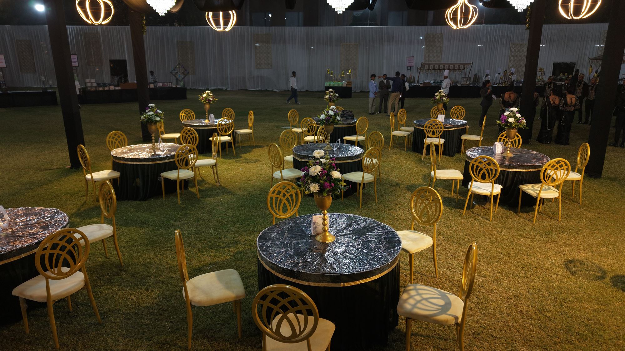 Black and gold gala dinner tables under wireframe lantern chandeliers on the lawn